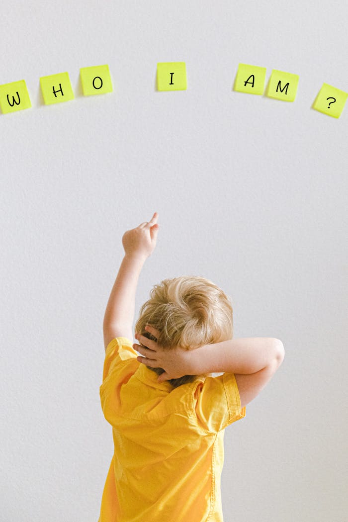 Blond child in yellow shirt pointing at words WHO I AM? on sticky notes on a wall.